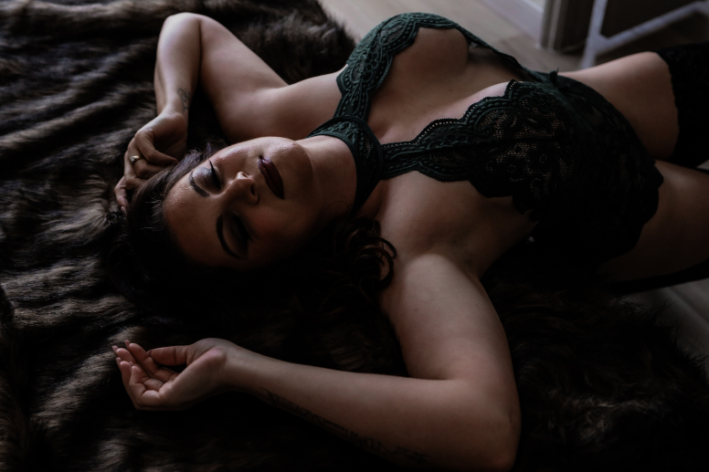 A woman in dark lace lingerie is lying on a fur blanket with her eyes closed and arms above her head, creating a relaxed and sensual atmosphere.
