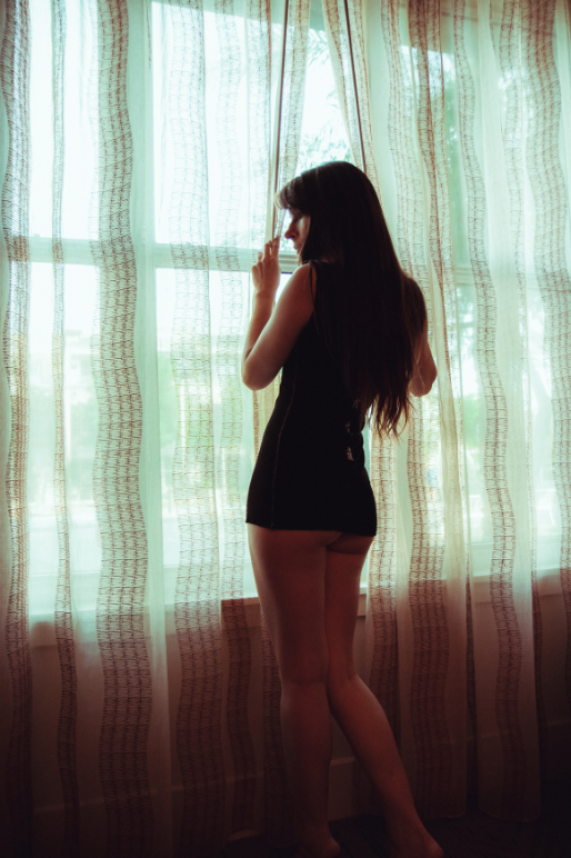 A woman in a black outfit stands barefoot by a window with sheer curtains, looking outside and holding the curtain with one hand. Soft daylight filters through the window.