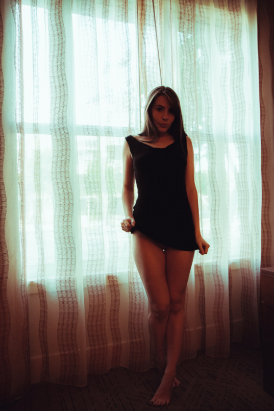 A woman in a short black dress stands barefoot in front of a sunlit window with sheer curtains, looking towards the camera.