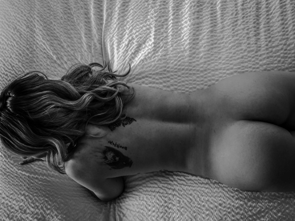 A black-and-white, high-angle shot of a person with blonde wavy hair lying face-down on a textured bedspread. They are nude, revealing tattoos on their upper back, and their head is turned slightly away from the camera.