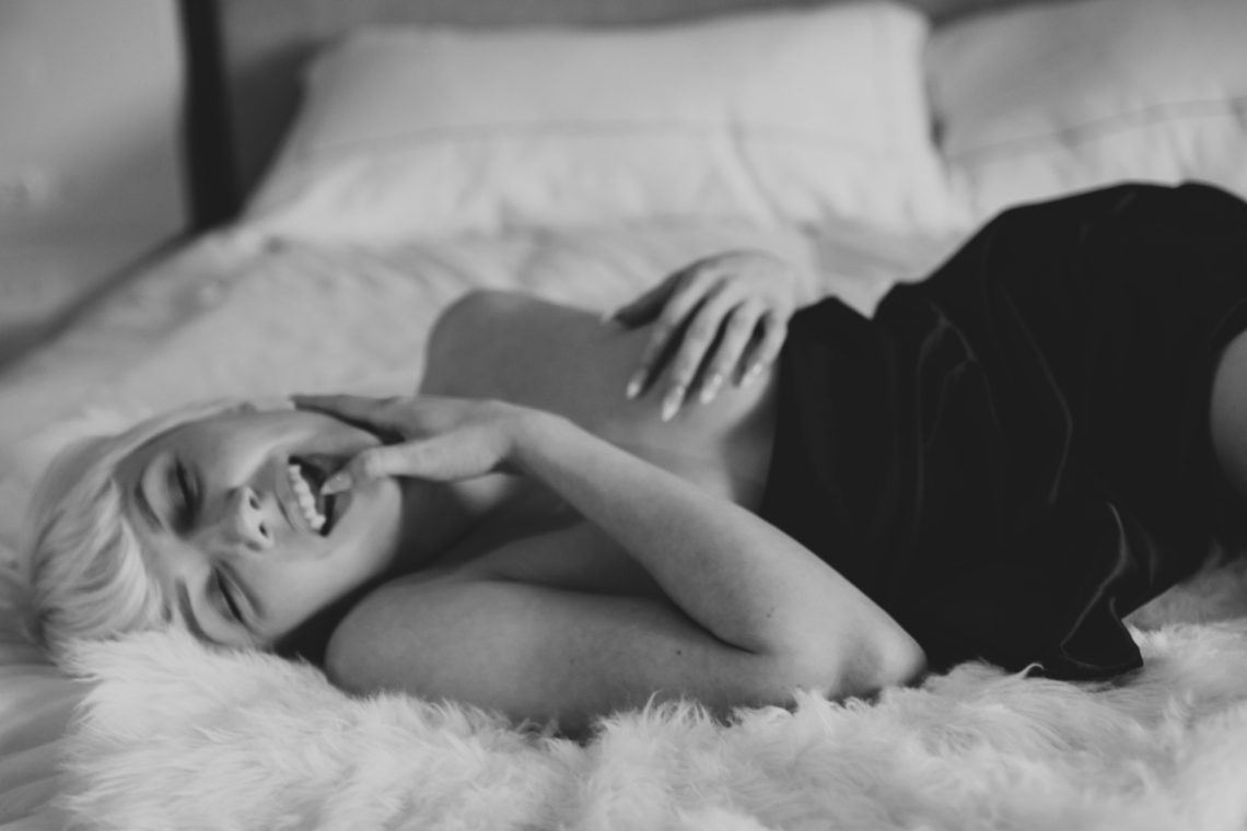 A person with short light hair lies on a bed, smiling and laughing with one hand touching their face. The photo is in black and white and has a relaxed, playful mood.