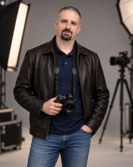 A man in a leather jacket and polo shirt holding a camera in a studio.