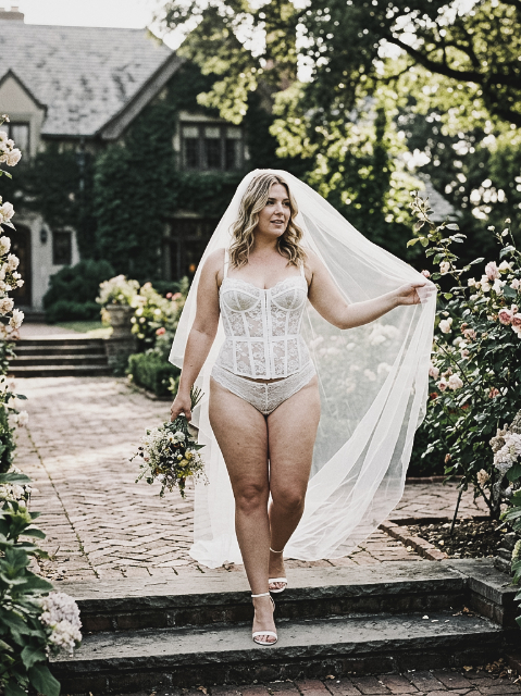 A bride wearing a lace corset and matching bottoms with a long veil walks down stone steps in a garden, holding a bouquet of wildflowers.