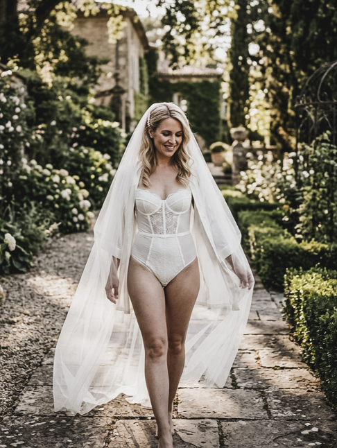 A smiling woman wearing a white bridal bodysuit and veil walks along a garden path.