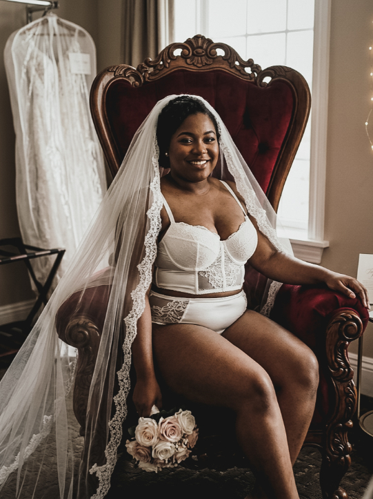 A smiling bride in white bridal lingerie and a lace-trimmed veil sits in a red velvet chair holding a small bouquet of roses.