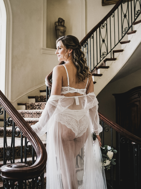 A person standing on a staircase wearing a white lace lingerie set and a sheer robe, holding a small bouquet of white flowers.