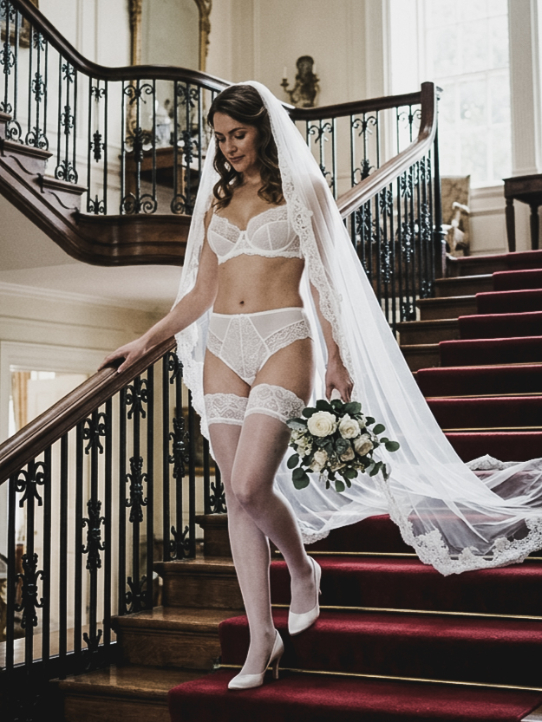 A bride in white lace lingerie, veil, and stockings walks down a carpeted staircase carrying a small bouquet.