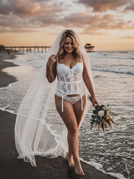 A smiling woman in a white lace bustier, matching bottoms, and a long veil walks along the shore holding a bridal bouquet.