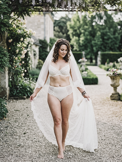 A smiling bride wearing a white lace lingerie set and a long, floor-length veil walks along a gravel path in a garden.