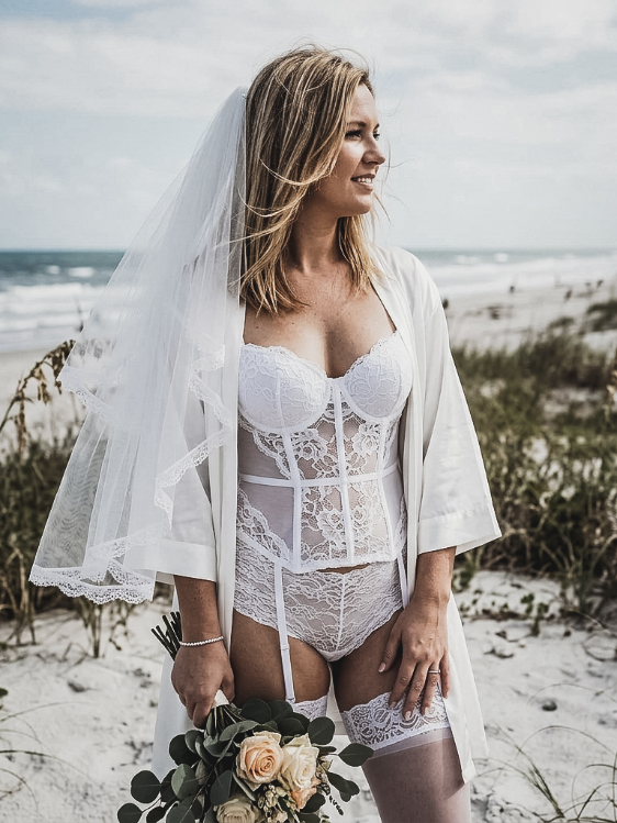 A bride wearing a white lace corset, matching lingerie, and a robe, holding a bouquet on a beach.