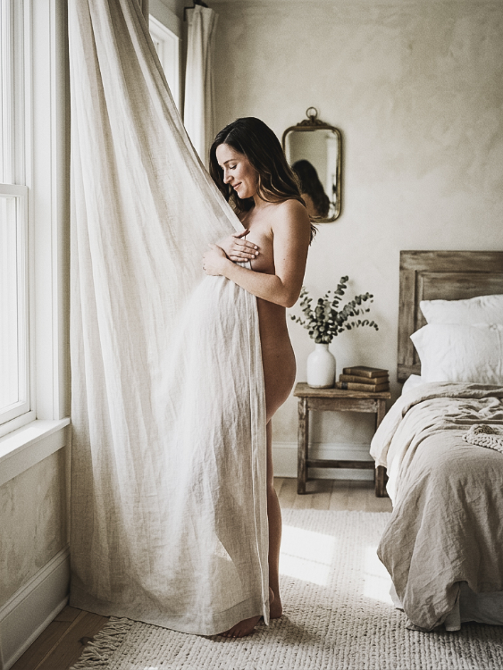 A pregnant woman stands by a window holding a curtain to her body in a sunlit bedroom.