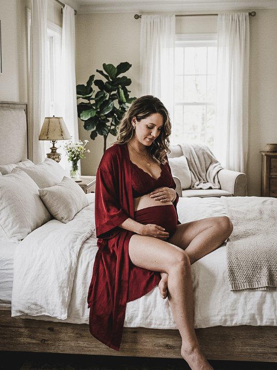 A pregnant woman in a deep red robe and lace lingerie sits on a bed, resting her hands on her belly.