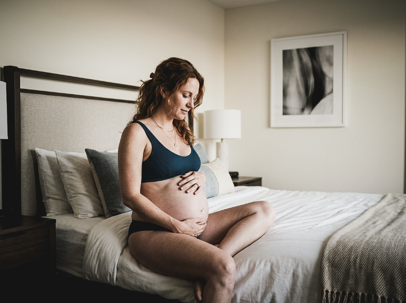A pregnant woman sits cross-legged on a bed, gently holding her belly with both hands.