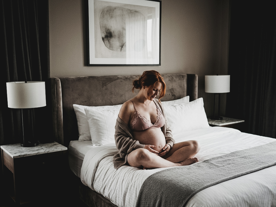 A pregnant woman in a bra and cardigan sitting cross-legged on a bed, looking down at her belly.