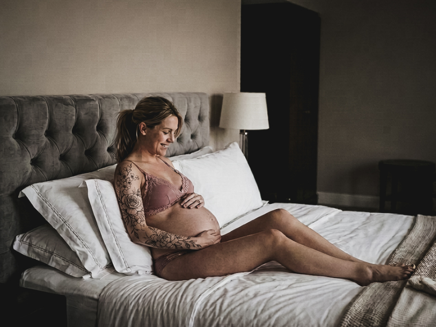 A pregnant woman with a floral tattoo on her arm sits on a bed, smiling down at her baby bump.